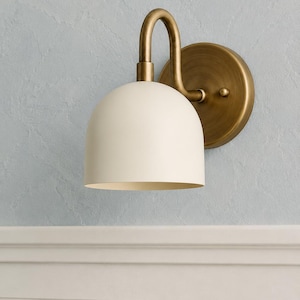 May include: A wall-mounted light fitting featuring a matte off-white dome-shaped shade attached to a curved arm and a circular backplate, both finished in a brushed antique brass tone, providing a downward-facing light source for interior spaces.