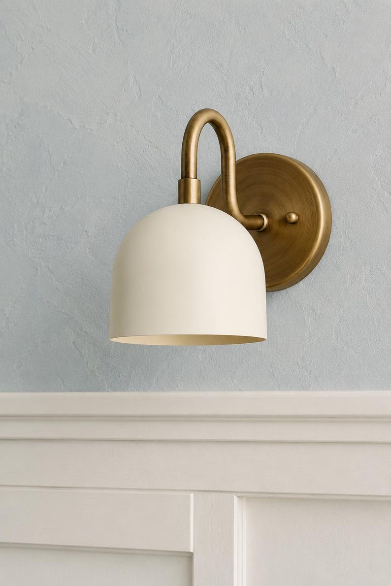 May include: A wall-mounted light fitting featuring a matte off-white dome-shaped shade attached to a curved arm and a circular backplate, both finished in a brushed antique brass tone, providing a downward-facing light source for interior spaces.
