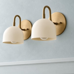 May include: Two wall-mounted light fittings featuring matte cream-coloured dome shades and curved brass arms. Each lamp is attached to a circular brass wall plate, with a single light bulb visible inside the downward-facing dome shade.