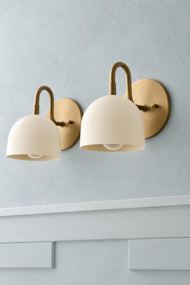 May include: Two wall-mounted light fittings featuring matte cream-coloured dome shades and curved brass arms. Each lamp is attached to a circular brass wall plate, with a single light bulb visible inside the downward-facing dome shade.