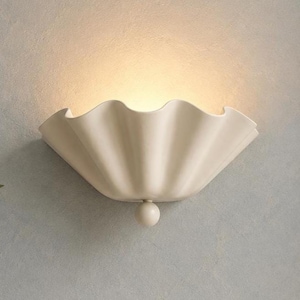 Könnte beinhalten: Eine cremefarbene Wandleuchte mit einem breiten, gewellten, muschelartigen Schirm mit glatter, matter Oberfläche. Die Leuchte strahlt ein warmes, sanftes Licht aus ihrer Mitte aus und besitzt einen kleinen, kugelförmigen Zierknopf am unteren Sockel.