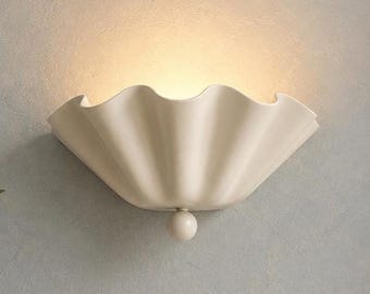 May include: A cream-colored wall sconce featuring a wide, scalloped, shell-like shade with a smooth matte finish. The fixture emits a warm, soft glow from its center and includes a small, spherical decorative knob at the bottom base.
