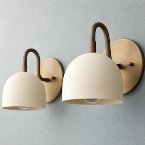 May include: Two wall sconces featuring matte cream-coloured dome shades and curved arms attached to circular brass backplates. The fixtures have a smooth finish and a simple, rounded design, with the light bulbs visible inside the dome shades.
