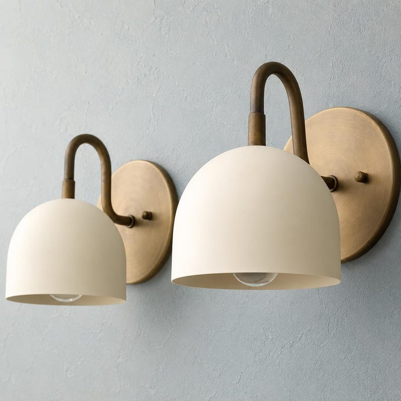 May include: Two wall sconces featuring matte cream-coloured dome shades and curved arms attached to circular brass backplates. The fixtures have a smooth finish and a simple, rounded design, with the light bulbs visible inside the dome shades.
