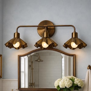 Puede incluir: Un aplique de pared para baño de tres luces con acabado en latón cepillado. La lámpara cuenta con un soporte circular y una barra horizontal que sostiene tres pantallas de metal en forma de campana con bordes ondulados y bombillas brillantes.