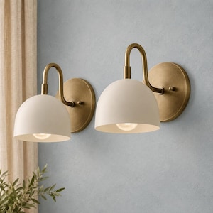 May include: A pair of wall sconces featuring matte off-white dome-shaped shades attached to curved arms and circular backplates finished in a brushed antique brass tone. Each fixture includes a single light bulb centred within the dome shade.