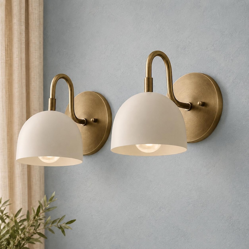 May include: A pair of wall sconces featuring matte off-white dome-shaped shades attached to curved arms and circular backplates finished in a brushed antique brass tone. Each fixture includes a single light bulb centred within the dome shade.