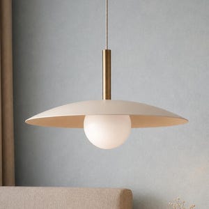 May include: A pendant light fixture featuring a wide, shallow, matte beige saucer-shaped shade suspended by a thin cord. A cylindrical brass-toned metal stem connects the shade to a central, spherical, frosted white glass globe light bulb.