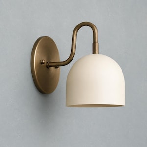 May include: A wall-mounted light fitting featuring a circular antique brass backplate and a curved arm that supports a smooth, dome-shaped off-white shade. The metal arm connects to the top of the shade, directing light downwards from the fitting.