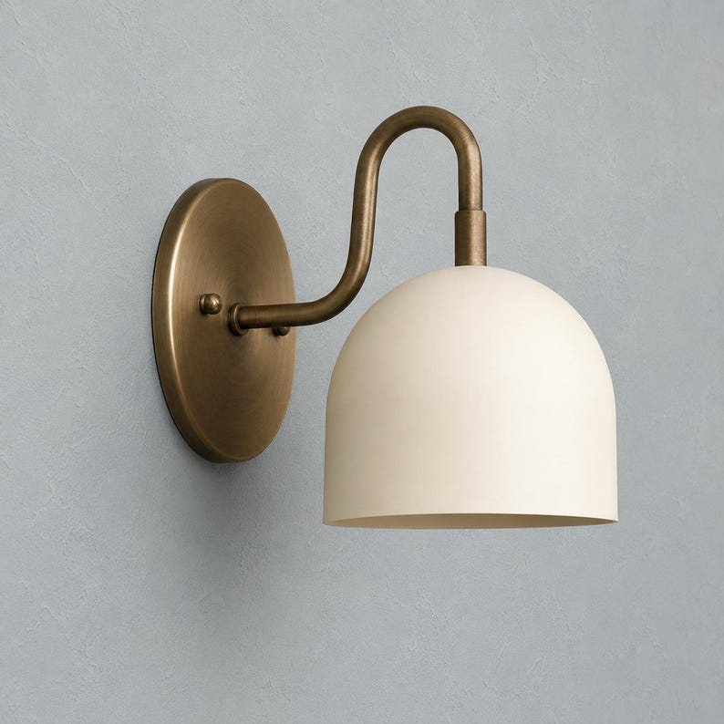 May include: A wall-mounted light fitting featuring a circular antique brass backplate and a curved arm that supports a smooth, dome-shaped off-white shade. The metal arm connects to the top of the shade, directing light downwards from the fitting.
