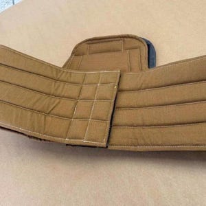 May include: A tan, padded support belt featuring horizontal and vertical stitched lines across the fabric panels. The belt has a central back panel and two side wings that wrap around, constructed with reinforced seams and a structured design.