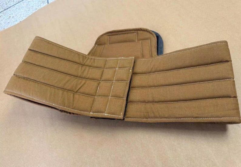 May include: A tan, padded support belt featuring horizontal and vertical stitched lines across the fabric panels. The belt has a central back panel and two side wings that wrap around, constructed with reinforced seams and a structured design.