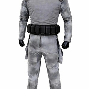 May include: A grey jumpsuit with a mottled, weathered texture, featuring a high collar, long sleeves, and dark shoulder patches. A black utility belt with multiple rectangular pouches is worn around the waist. The garment includes large thigh pockets.