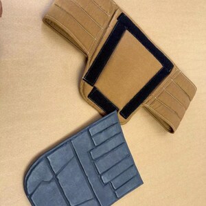 May include: A tan fabric wrap with black hook-and-loop fastener strips and a separate dark gray, rectangular, multi-sectioned padded insert. The tan wrap features horizontal stitching lines and a central rectangular area framed by black fasteners.