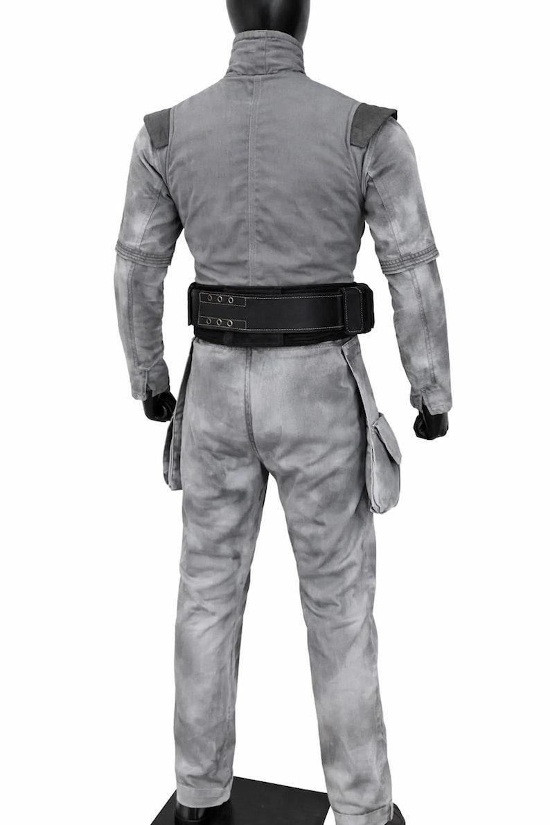 May include: A light gray jumpsuit with a mottled, weathered texture, featuring a high collar, long sleeves, and two side pockets on the thighs. A wide black utility belt with visible stitching and metal eyelets is fastened around the waist.