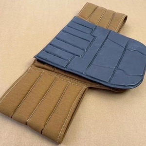 May include: A tan fabric belt featuring vertical quilted stitching and a dark gray, rectangular, padded panel attached to the center. The panel has a segmented, grid-like surface texture with distinct horizontal and vertical lines.