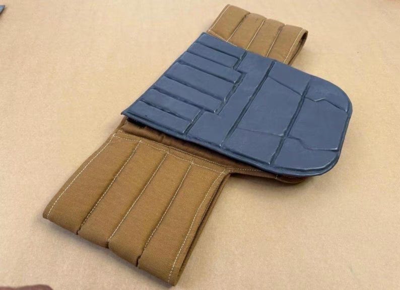 May include: A tan fabric belt featuring vertical quilted stitching and a dark gray, rectangular, padded panel attached to the center. The panel has a segmented, grid-like surface texture with distinct horizontal and vertical lines.