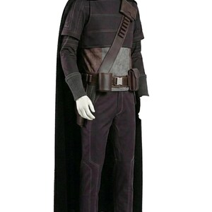 K&ouml;nnte beinhalten: Ein dunkelvioletter Anzug mit einem schwarzen Umhang. Das Outfit umfasst ein lang&auml;rmeliges Hemd, eine Hose und einen G&uuml;rtel mit Taschen. Das Kost&uuml;m hat ein futuristisches Design mit einem braunen Lederriemen &uuml;ber der Brust.