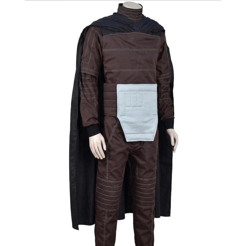May include: A dark brown jumpsuit featuring a high collar, long sleeves with white stitching, and ribbed detailing on the legs. A light gray rectangular panel is attached to the waist. A long black cape is draped over the shoulders and back.