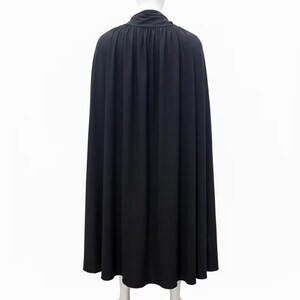 May include: A floor-length black cape featuring a high, gathered collar and a full, flowing silhouette. The garment is displayed on a mannequin, showcasing vertical pleats that extend from the neckline down to the hem, creating a draped effect.