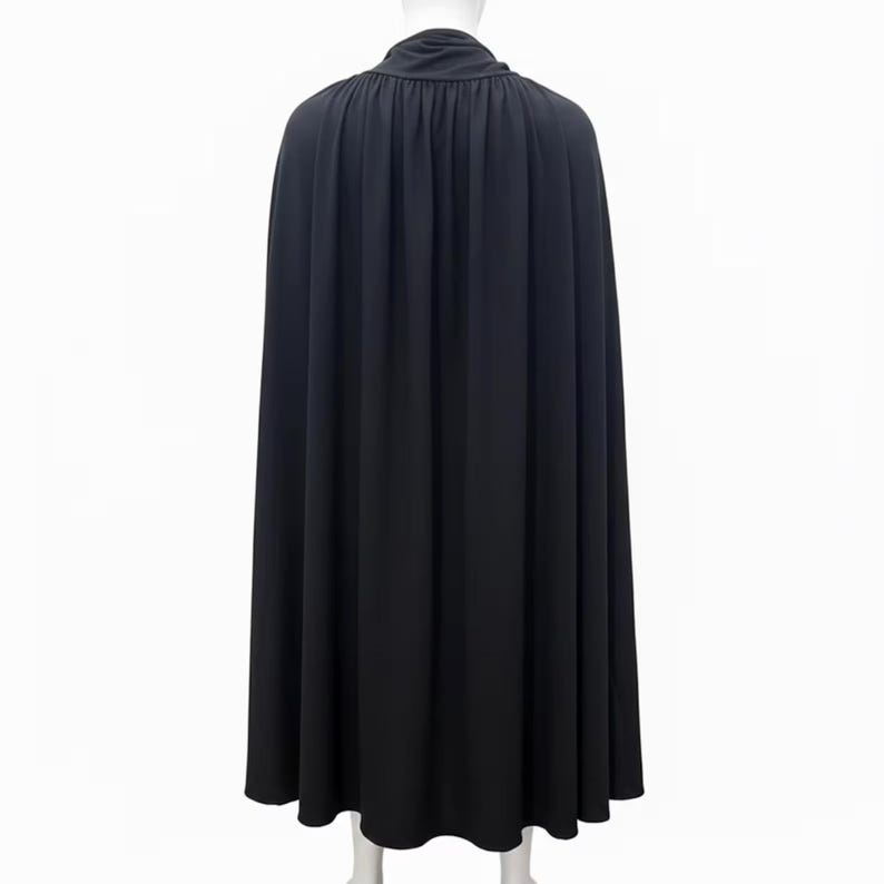 May include: A floor-length black cape featuring a high, gathered collar and a full, flowing silhouette. The garment is displayed on a mannequin, showcasing vertical pleats that extend from the neckline down to the hem, creating a draped effect.