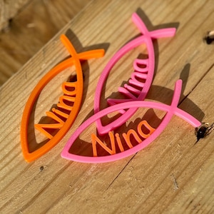 Könnte beinhalten: Drei fischförmige Ornamente in Orange und Rosa mit dem Namen "Nina" in Orange. Die Ornamente liegen auf einer Holzoberfläche.