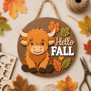 May include: A circular wooden wall hanging featuring a layered cutout of a brown Highland cow, orange and green leaves, and a small heart. The text "Hello FALL" is displayed in white and tan script and block lettering with a rope hanger.