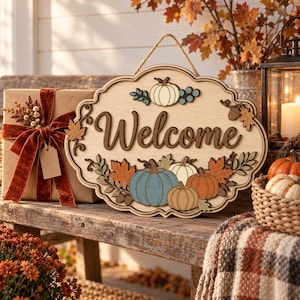 May include: A decorative wooden wall sign with a scalloped edge featuring the word Welcome in dark brown script. The sign is adorned with layered cutouts of pumpkins in blue, orange, and cream, along with autumn leaves and a jute rope hanger.