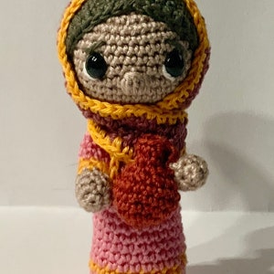 May include: A crocheted doll wearing a pink dress with yellow and red accents. The doll has brown yarn hair and large black eyes.