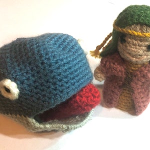 May include: A blue crocheted whale with a red tongue and a white eye, next to a brown and green crocheted figure wearing a yellow headband.