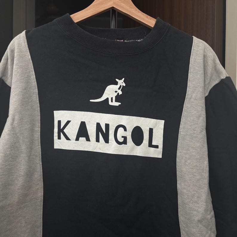 May include: Black and gray sweatshirt with the word "KANGOL" in a white rectangle. A white kangaroo logo is above the text. The sweatshirt is hanging on a wooden hanger.