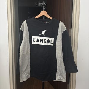May include: Black and gray long-sleeve shirt with the word "KANGOL" in a white rectangle. A white kangaroo logo is above the brand name. The shirt is hanging on a wooden hanger.