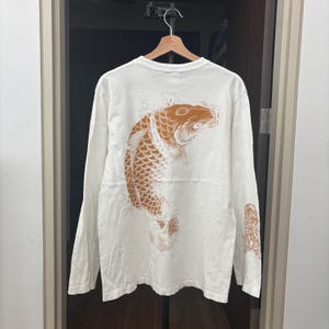 May include: Cream-coloured long-sleeved shirt with a large orange koi carp design on the back. The fish is detailed with scales and fins, and the shirt has a crew neck. The design extends to the right sleeve.