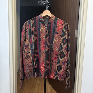 May include: A long-sleeve zip-up jacket featuring a dark base with a multi-colored pointillist pattern. The design includes vertical stripes, geometric diamond shapes, and floral motifs in shades of red, yellow, and purple. The jacket has a stand-up collar and a full-length front zipper closure.