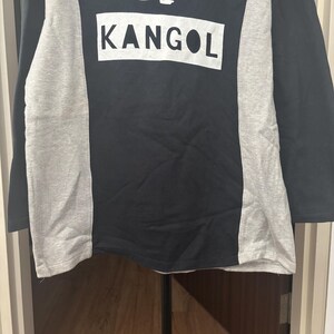 May include: Black long-sleeve shirt with gray side panels. The word "KANGOL" is printed in white block letters on the front of the shirt. The shirt is made of a soft, comfortable material.