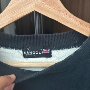 May include: Close-up of a black sweatshirt with a white interior, hanging on a wooden hanger. The sweatshirt features a black collar and a tag with the brand name "KANGOL" and a British flag.
