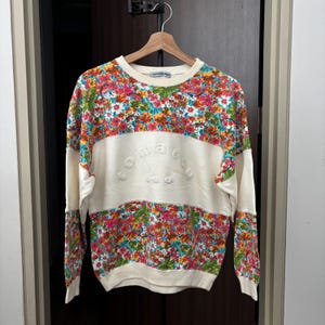 May include: A cream-colored sweatshirt with a floral pattern in pink, yellow, blue, and green. The sweatshirt has a horizontal cream-colored band across the chest with the embossed text "KOMATSU CLUB". The sleeves and upper back are also cream-colored.