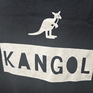May include: Black fabric with a white rectangular design that reads "KANGOL" in bold, sans-serif font. Above the text is a white kangaroo logo. The image is a close-up of the fabric.