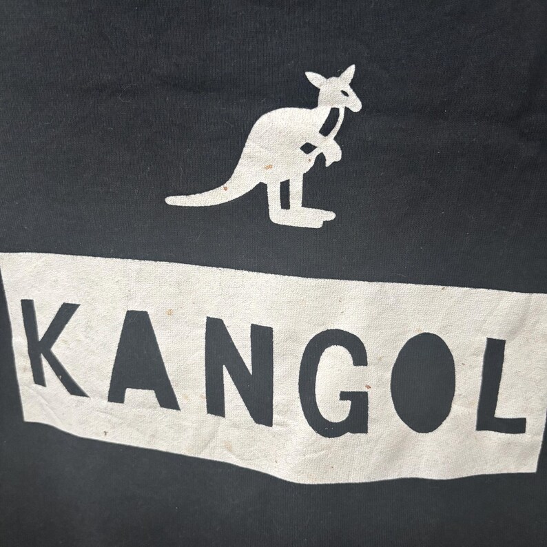 May include: Black fabric with a white rectangular design that reads "KANGOL" in bold, sans-serif font. Above the text is a white kangaroo logo. The image is a close-up of the fabric.