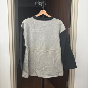 May include: A gray long-sleeved shirt with black sleeves and a black collar is hanging on a wooden hanger. The shirt is made of a soft material and is displayed against a dark background.