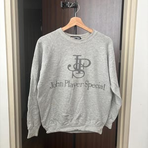 May include: A heather gray sweatshirt with the text "John Player Special" in a dark gray font. The sweatshirt has a crew neck and long sleeves. The logo is centered on the chest.