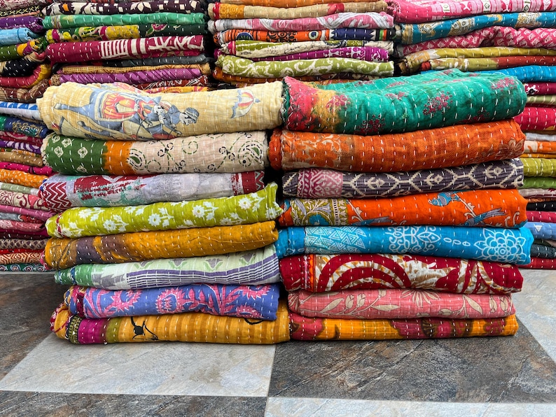 May include: Stacks of colorful, patterned quilts. The quilts display a variety of colors, including green, orange, blue, and pink. Some quilts feature intricate designs and patterns, while others have a more simple, solid appearance. The quilts are neatly folded and stacked.