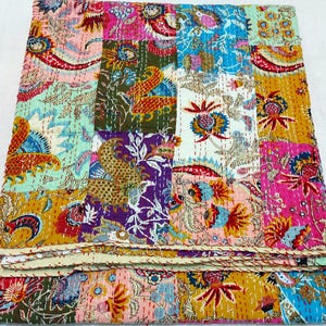 May include: A vibrant patchwork quilt featuring floral and paisley designs in pink, blue, yellow, and green. The quilt is constructed from numerous squares, sewn together with visible stitching. The folded edge is visible at the bottom.