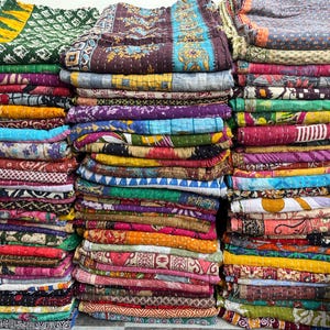 Puede incluir: Montones de colchas coloridas y estampadas. Las colchas muestran una variedad de colores, incluyendo verde, morado, naranja y azul. Cada colcha tiene un diseño único, con patrones florales y geométricos. Las colchas están cuidadosamente dobladas y apiladas.