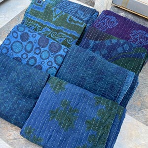 Könnte beinhalten: Eine Sammlung gefalteter, gesteppter Textilien in verschiedenen Blau- und Grüntönen. Die Stoffe weisen unterschiedliche Muster auf, darunter Kreise, florale Designs und geometrische Formen. Weiße Nähte sind auf den Stücken sichtbar und verleihen Textur und Details.