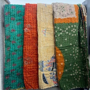 Puede incluir: Una exhibición de textiles acolchados coloridos. Los textiles son en tonos de verde, naranja y amarillo, con costuras y patrones intrincados. Algunos presentan diseños florales, mientras que otros tienen ilustraciones únicas. Los textiles están colgados al aire libre.