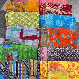 May include: A collection of colorful, folded kantha quilts. The quilts feature various patterns, including floral, geometric, and abstract designs. The color palette includes shades of blue, green, yellow, orange, and pink. The quilts are stacked, showcasing their stitched texture.