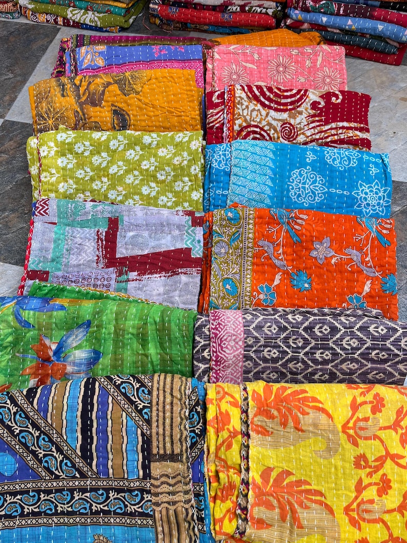 May include: A collection of colorful, folded kantha quilts. The quilts feature various patterns, including floral, geometric, and abstract designs. The color palette includes shades of blue, green, yellow, orange, and pink. The quilts are stacked, showcasing their stitched texture.