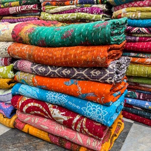 Puede incluir: Una pila de edredones enrollados de colores brillantes. Los edredones presentan diversos patrones y colores, incluyendo naranja, verde azulado, azul y rosa. Los edredones son de tela y tienen costuras visibles.