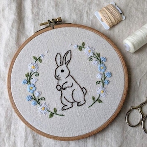 Puede incluir: Un bastidor de bordado hecho a mano con un diseño de conejo. El conejo está rodeado por una corona de flores azules y blancas. El bastidor de madera está sobre un fondo de lino neutro.